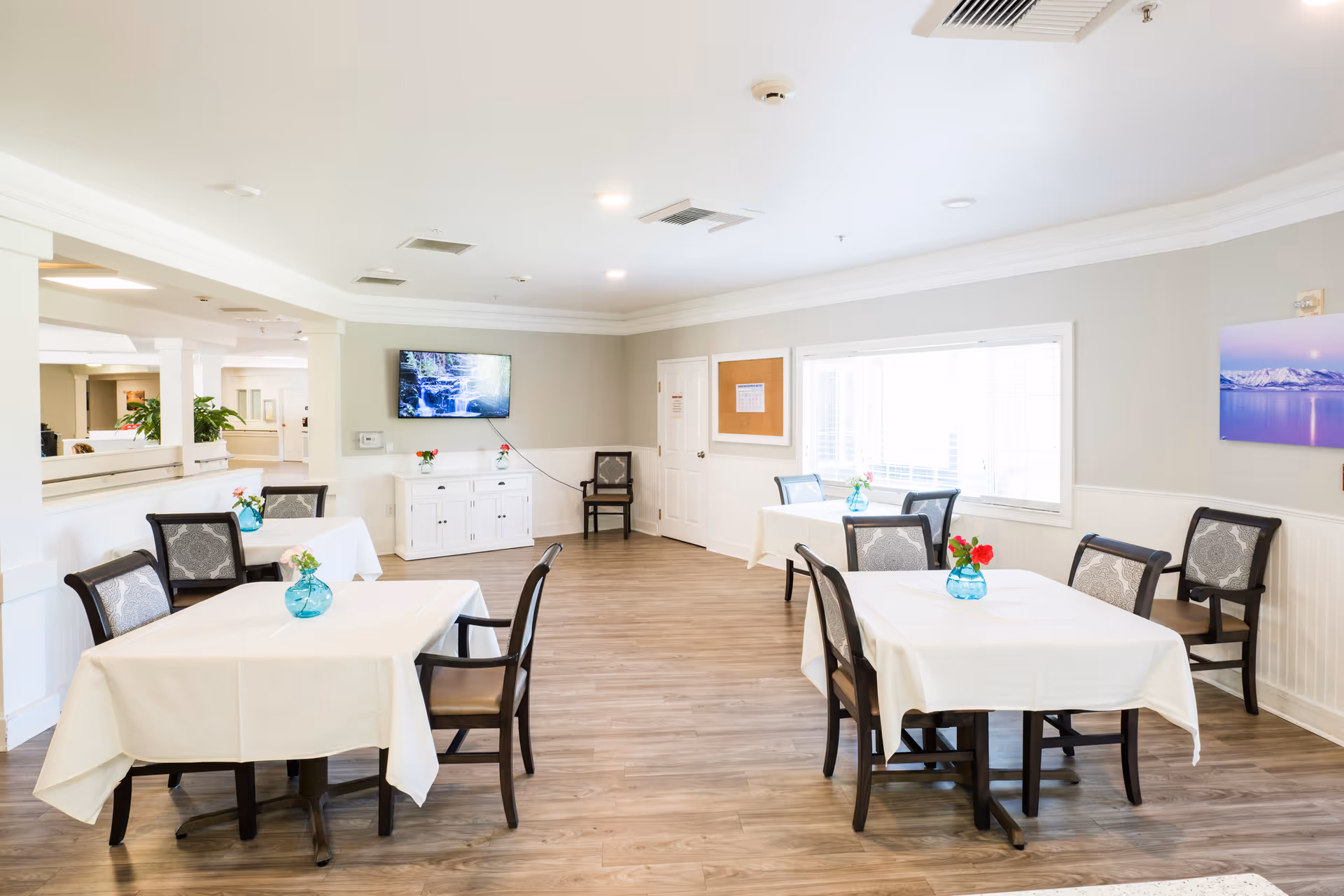 A bright and clean dining room with several tables covered in white tablecloths, each adorned with a small blue vase holding flowers. The room has wooden flooring, light-colored walls, a large window letting in natural light, a wall-mounted TV, and a white cabinet beneath it. There are multiple chairs around the tables and along the walls.