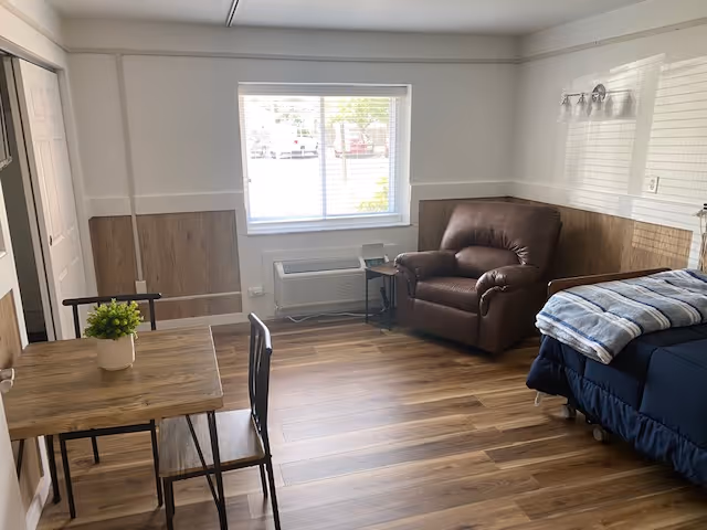 A senior living facility room with a wooden floor, a small wooden table with two chairs, a brown leather armchair, and a bed with a blue bedspread and a folded blanket on top. There is a window with blinds letting in natural light, and a small side table next to the armchair. The walls are white with wood paneling on the lower half.
