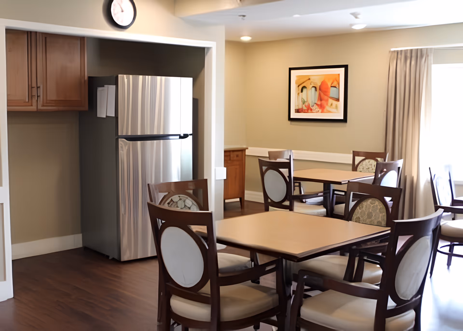 A dining area with several wooden tables and chairs with cushioned seats and backs. There is a stainless steel refrigerator in a small alcove with wooden cabinets above it. A clock is mounted above the alcove, and a framed artwork hangs on the wall. Natural light enters through a window with beige curtains.