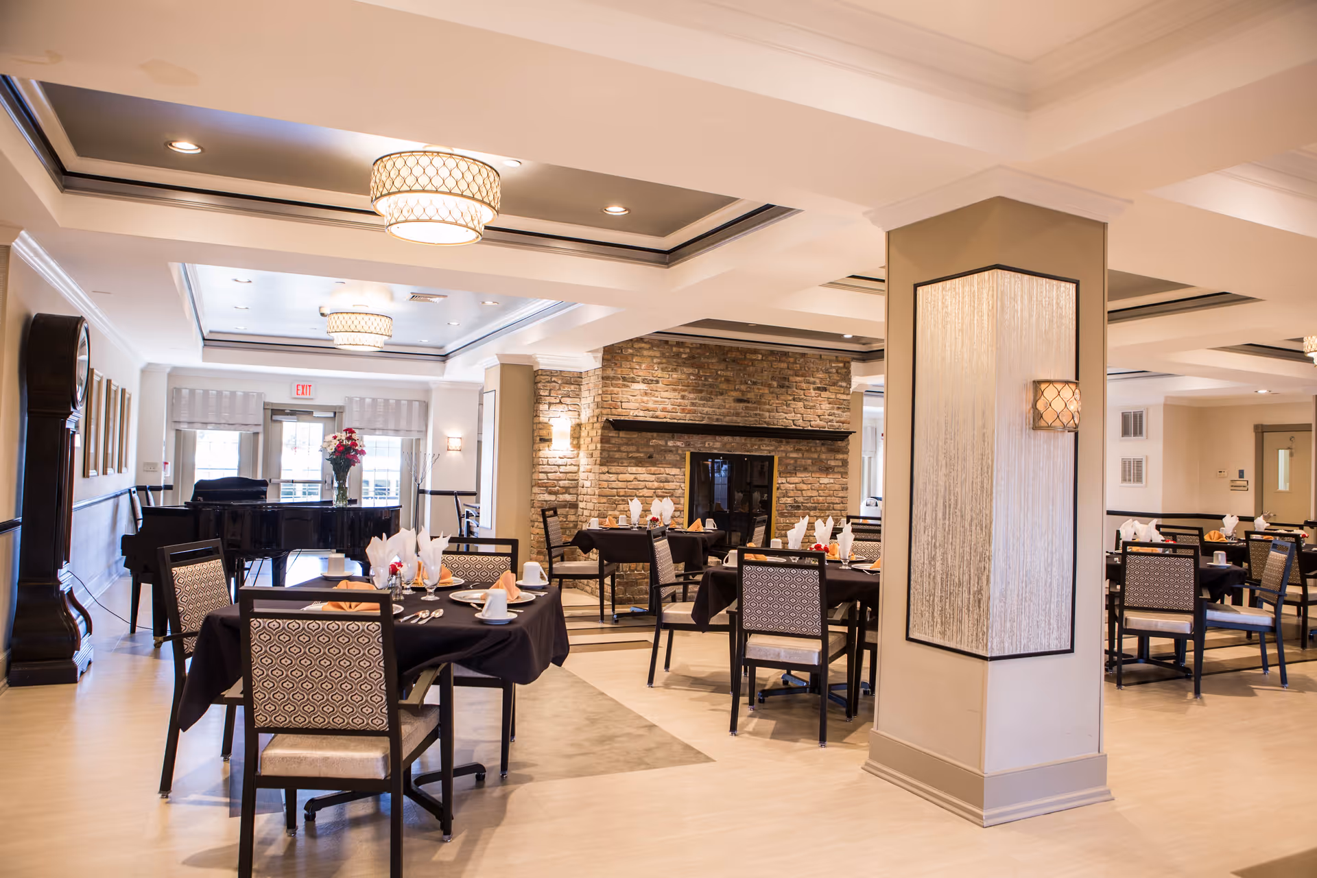 A spacious and elegant dining room with multiple tables set with black tablecloths, napkins, cups, and silverware. The room features a brick fireplace, a grand piano, a tall grandfather clock, and modern ceiling lights. The walls are decorated with framed pictures and a large floral arrangement is visible near the piano.