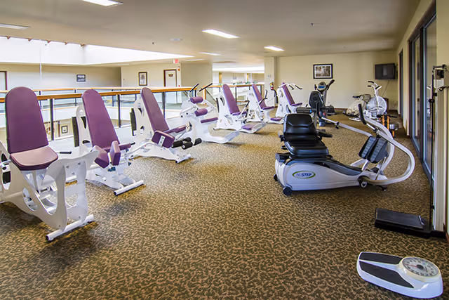 A bright indoor fitness area with multiple purple-and-white exercise machines and a recumbent bike on a carpeted floor overlooking an interior atrium.
