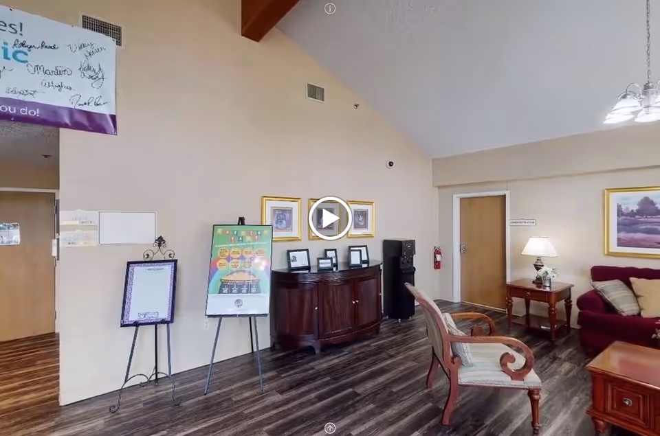 Interior view of a senior living facility common area with wooden flooring, a wooden sideboard with framed pictures, a water cooler, a couple of chairs, a sofa with cushions, a side table with a lamp, and framed artwork on the walls. There are also informational boards on stands and a door labeled 'COMMUNICATION'.