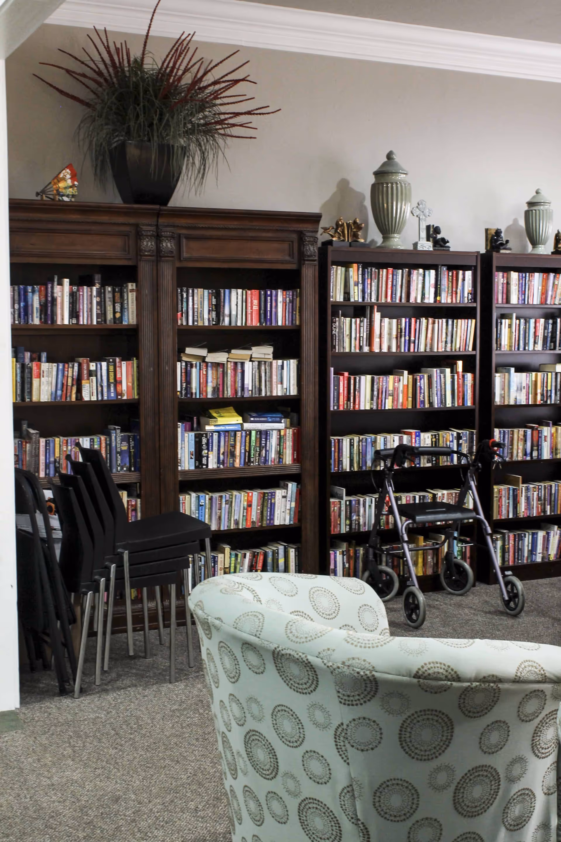 Interior of a senior living facility library or reading area with multiple bookshelves filled with books, a patterned armchair in the foreground, a stack of black chairs, and a walker positioned near the bookshelves. Decorative items and plants are placed on top of the bookshelves.