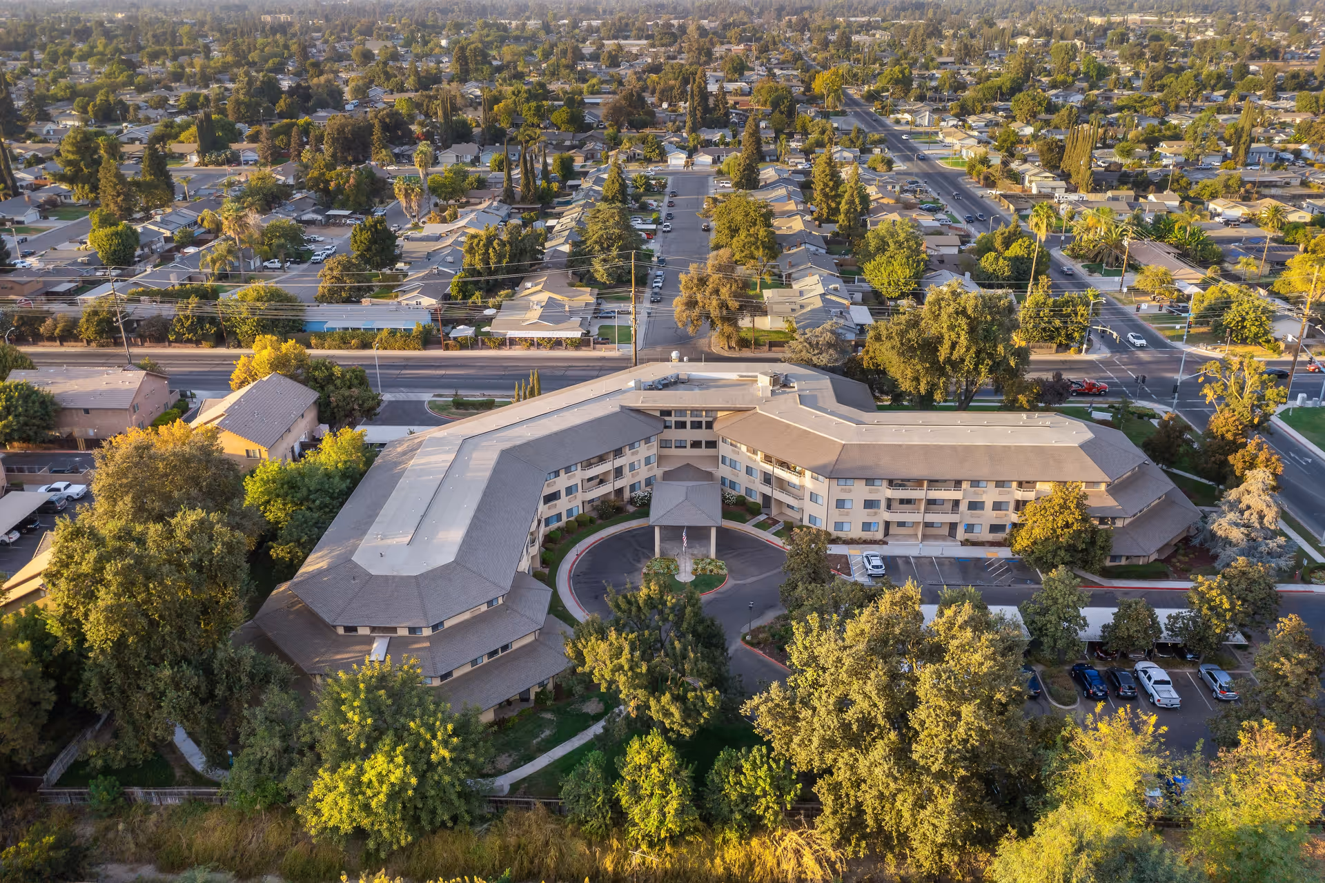 Aerial view of Walnut Park - A Provincial Senior Living Community, showing a large, U-shaped building surrounded by trees and greenery. The building has multiple floors and a covered entrance with a circular driveway. The surrounding neighborhood includes residential houses, streets, and parked cars.