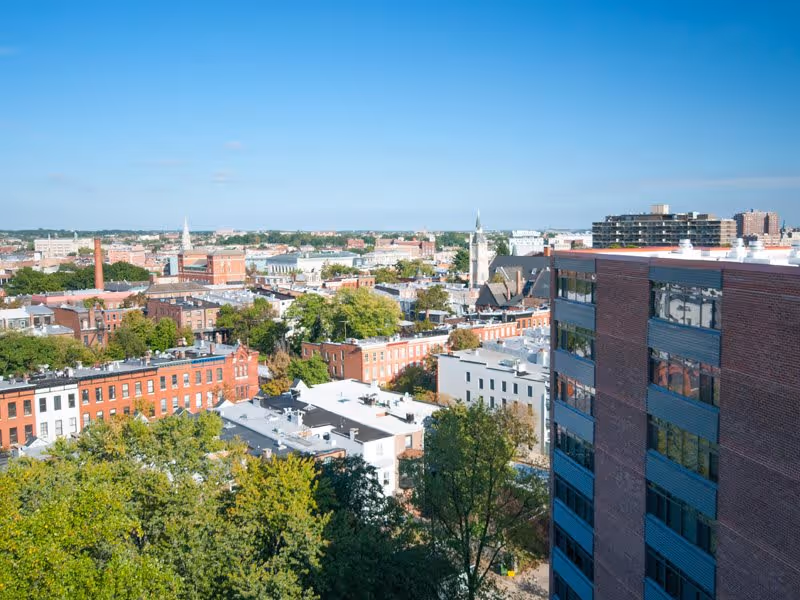 High-angle exterior view of a city neighborhood with rows of brick townhouses, trees, and a taller brick apartment building on the right under a clear blue sky.