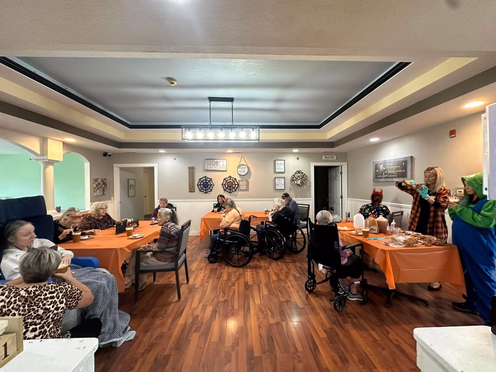 Seniors seated around orange-covered tables in a communal dining room while staff serve food.