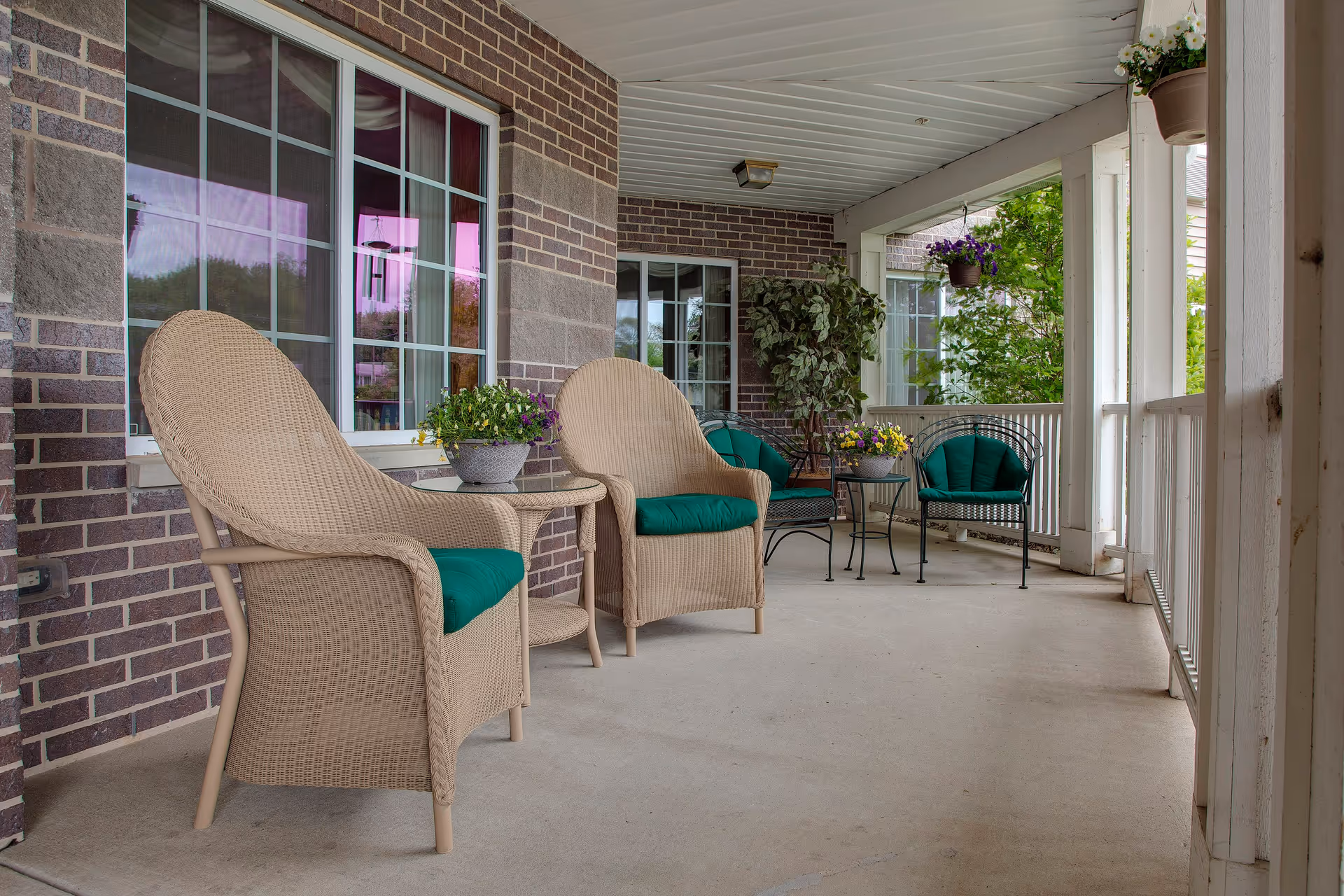 Covered porch with wicker chairs topped with green cushions, small tables and potted plants along a brick exterior.
