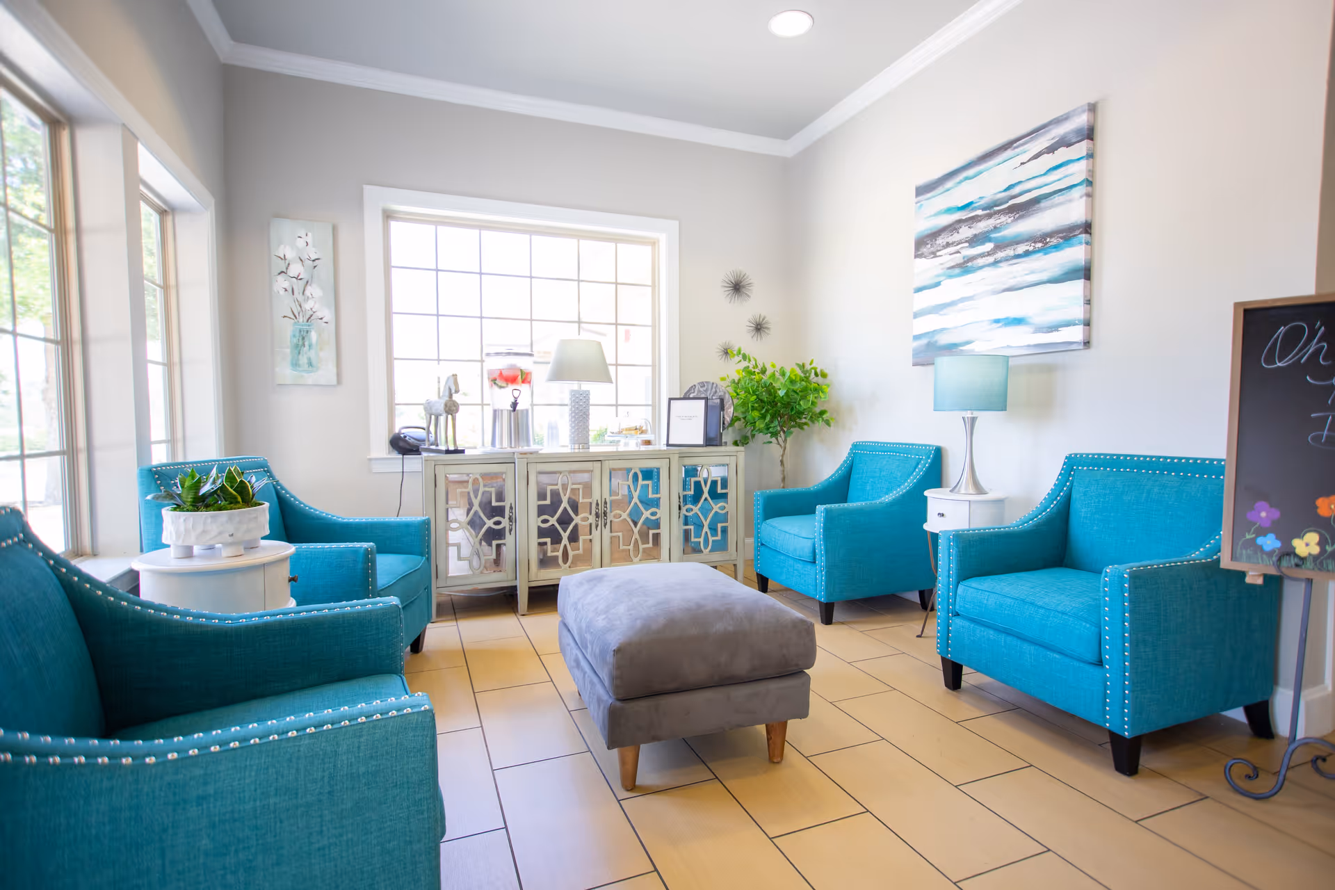 A bright and inviting sitting area with four teal armchairs arranged around a gray ottoman. The room features large windows letting in natural light, a decorative cabinet with a lamp and plants on top, and modern wall art. The floor is tiled in a light wood pattern.