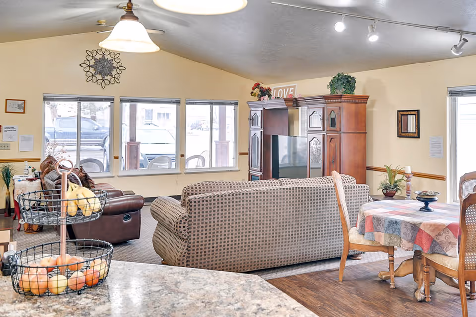 A cozy living room area in an assisted living facility with a patterned sofa and a brown leather recliner facing a wooden entertainment center with a TV. There is a round dining table with a colorful tablecloth and wooden chairs to the right. Large windows let in natural light, and a two-tiered fruit basket is visible on a granite countertop in the foreground.