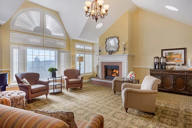 A cozy living room with a fireplace, several armchairs, a large window with white shutters, a chandelier, and a sideboard with coffee dispensers and framed artwork.