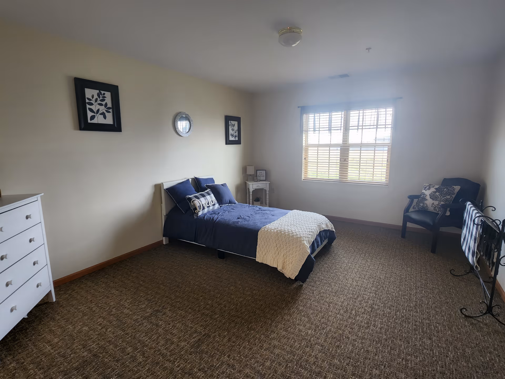 A simple bedroom with a single bed made with navy blue bedding and a white knitted throw. The room has beige walls with two framed pictures and a round mirror above the bed. There is a white dresser on the left side and a small white nightstand with a lamp and decor next to the bed. A window with blinds lets in natural light on the right wall. A black chair with a decorative pillow and a black metal towel rack with a striped towel are also visible in the room.