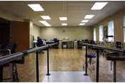 Rehabilitation therapy room with parallel gait bars, stools, exercise equipment and desks under fluorescent ceiling lights.