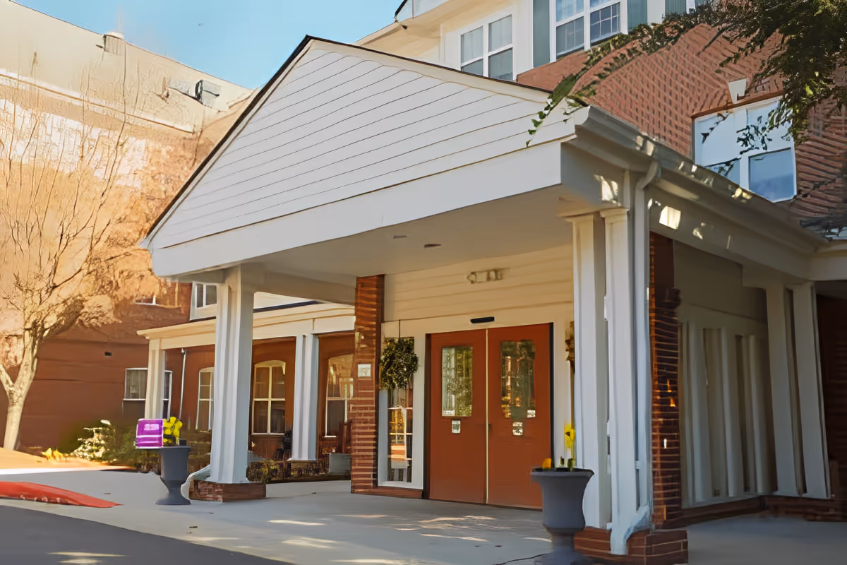 Entrance of a senior living facility with a covered drop-off area supported by white columns, double red doors, and potted plants on either side. The building has brick and white siding exterior with windows above the entrance.