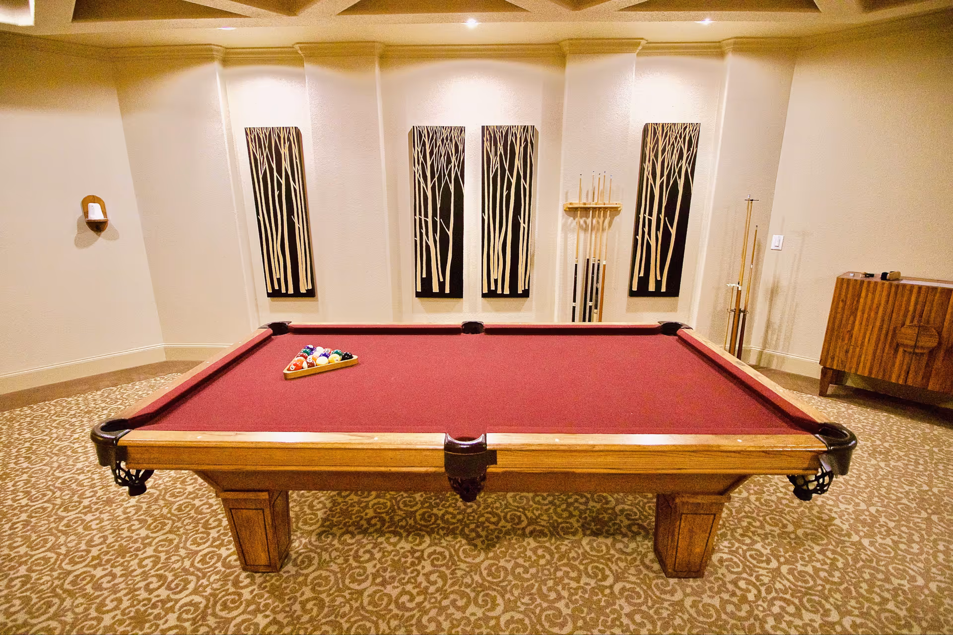 A room with a red felt pool table centered on a patterned carpet. The pool table has a wooden frame and a rack of billiard balls arranged on it. Behind the table, there are four vertical wall art pieces depicting stylized tree trunks in black and white. To the right, there is a wooden cabinet and a rack holding pool cues.