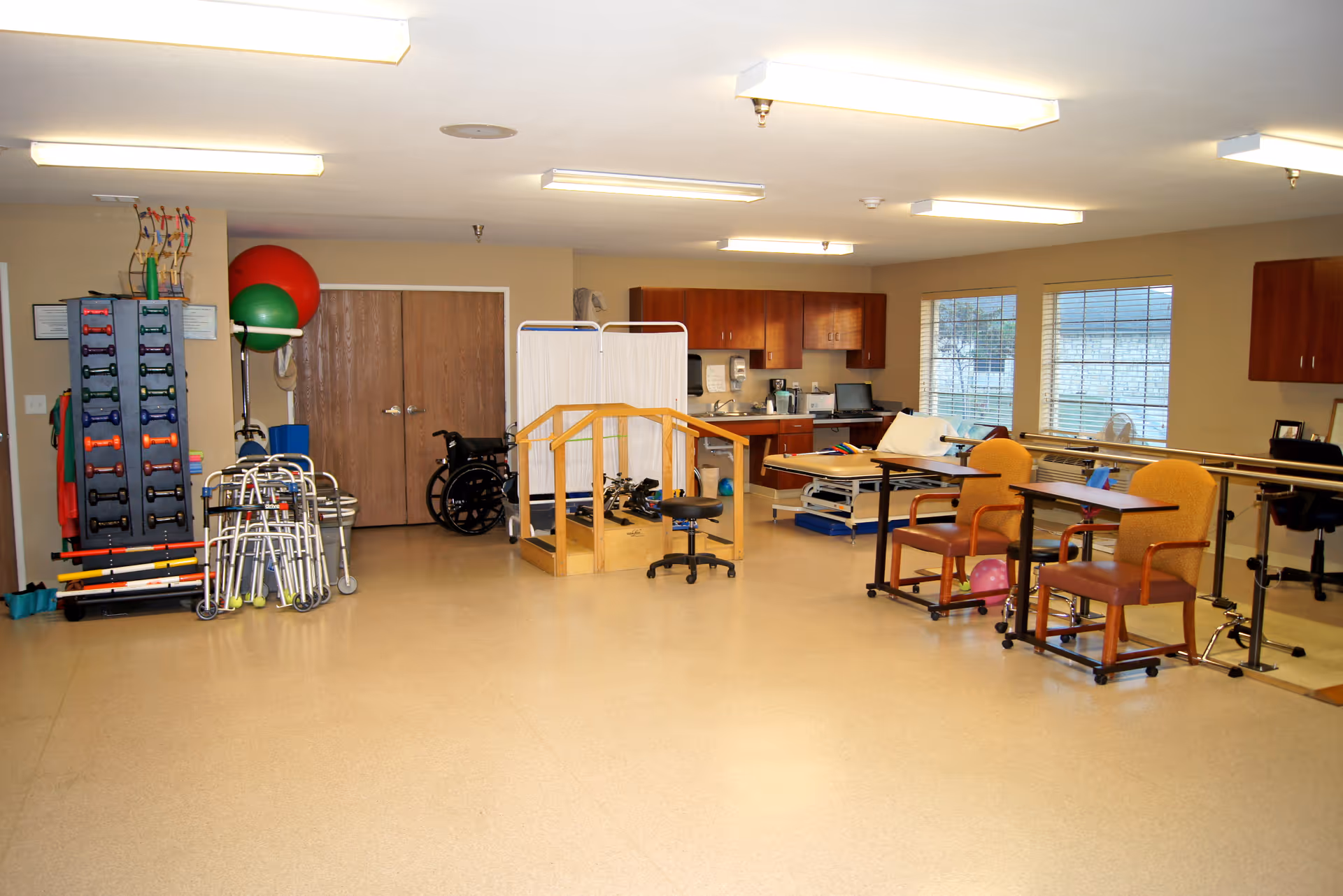 A rehabilitation room with exercise equipment including dumbbells, walkers, therapy balls, parallel bars, and chairs with tables. The room has beige walls, large windows, wooden cabinets, and bright overhead lighting.