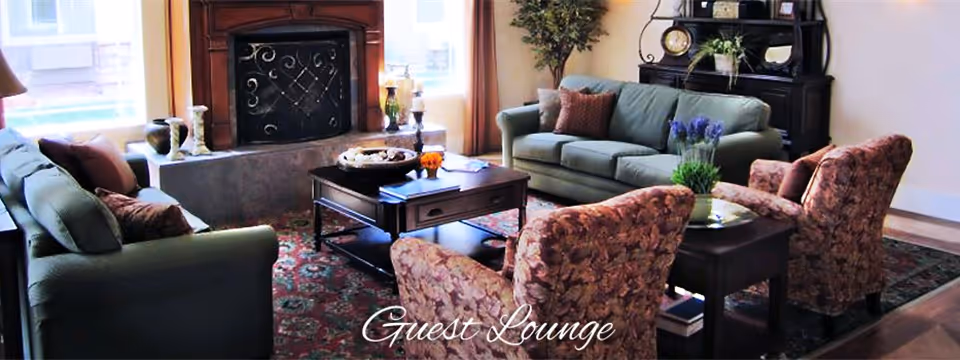A cozy guest lounge with two green sofas, two floral armchairs, a wooden coffee table, a side table with a plant, a fireplace with decorative items on the mantel, and a patterned rug on the floor.