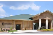 Exterior view of Arbor Springs Health And Rehab Center building with a green roof, beige brick walls, and a covered entrance under a blue sky with some clouds.