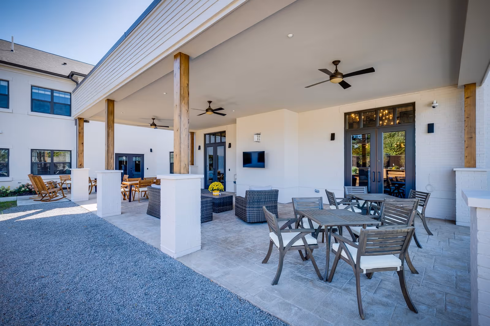 Covered outdoor patio area with wooden columns, ceiling fans, multiple seating arrangements including wooden tables with chairs and wicker lounge chairs around a coffee table, adjacent to a white brick building with large windows and glass doors.