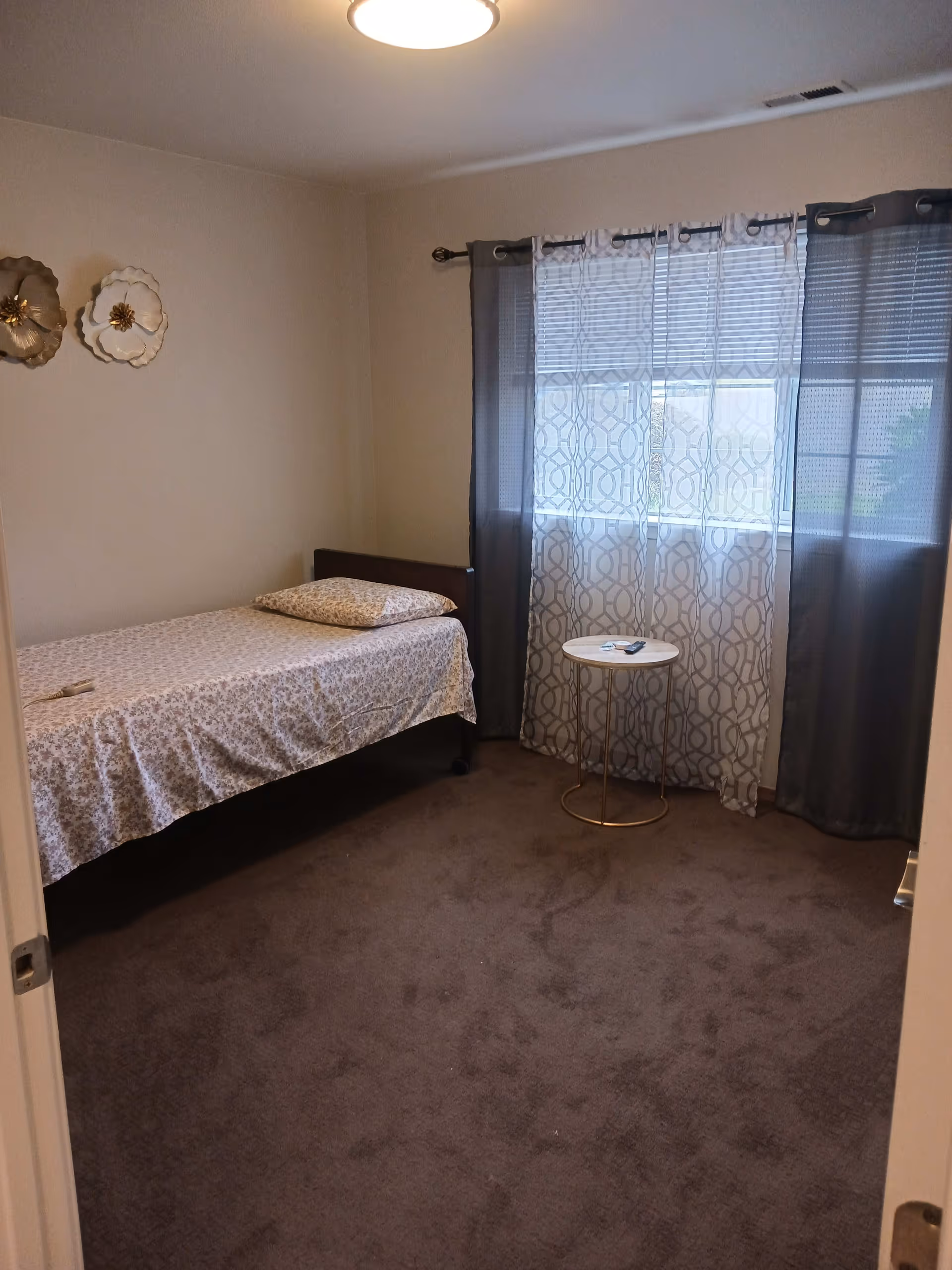 A simple bedroom with a single bed covered in floral bedding, a pillow, and a small round side table near a window with sheer patterned curtains and darker outer curtains. The walls are light-colored with two decorative wall hangings, and the floor is carpeted in a dark color.