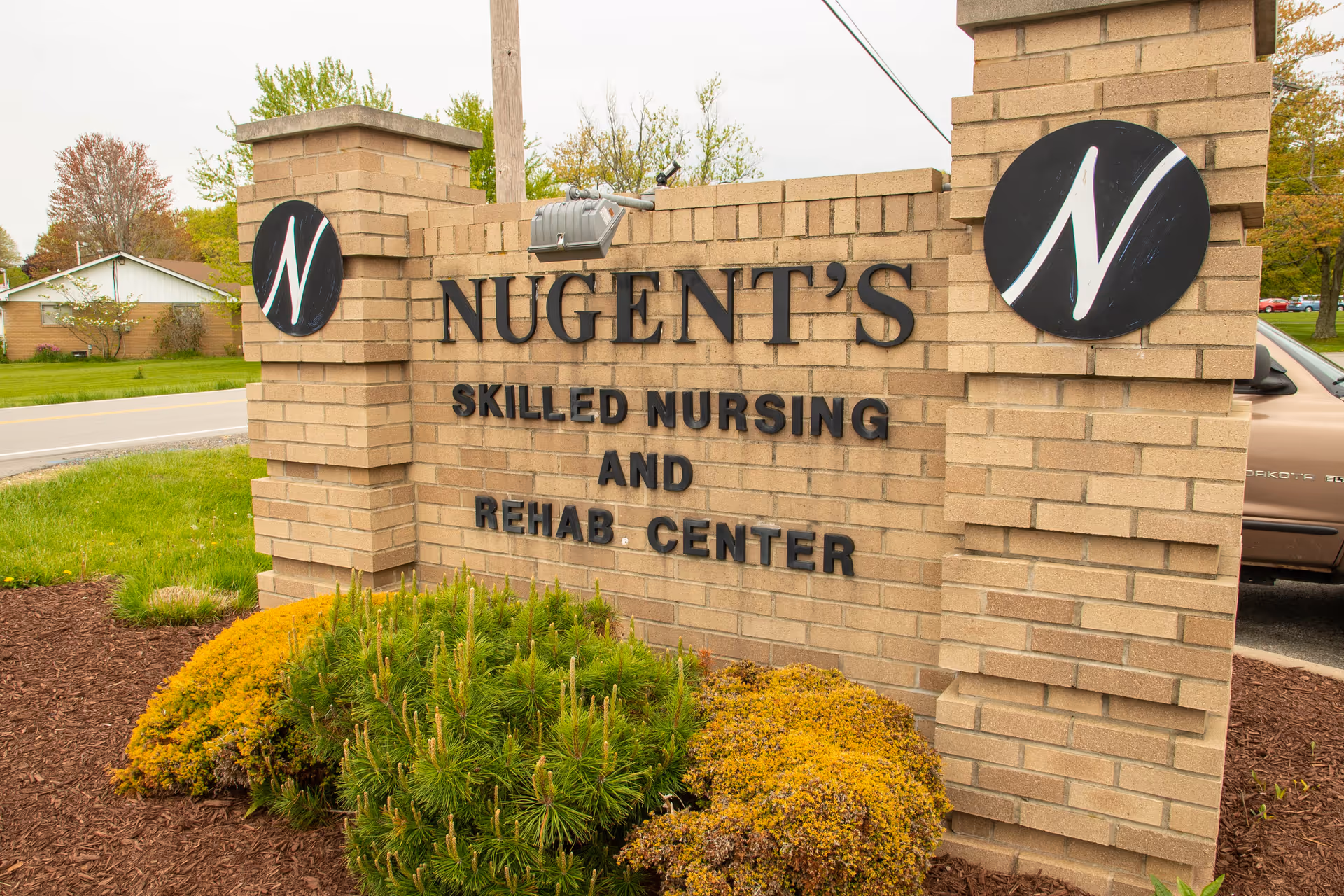 Brick sign for Nugent's Skilled Nursing and Rehab Center with landscaping in front and a road and houses in the background.
