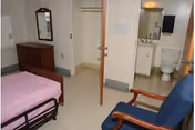 Small bedroom with a single hospital-style bed, dresser and mirror, closet, armchair, and an open door showing an attached bathroom.