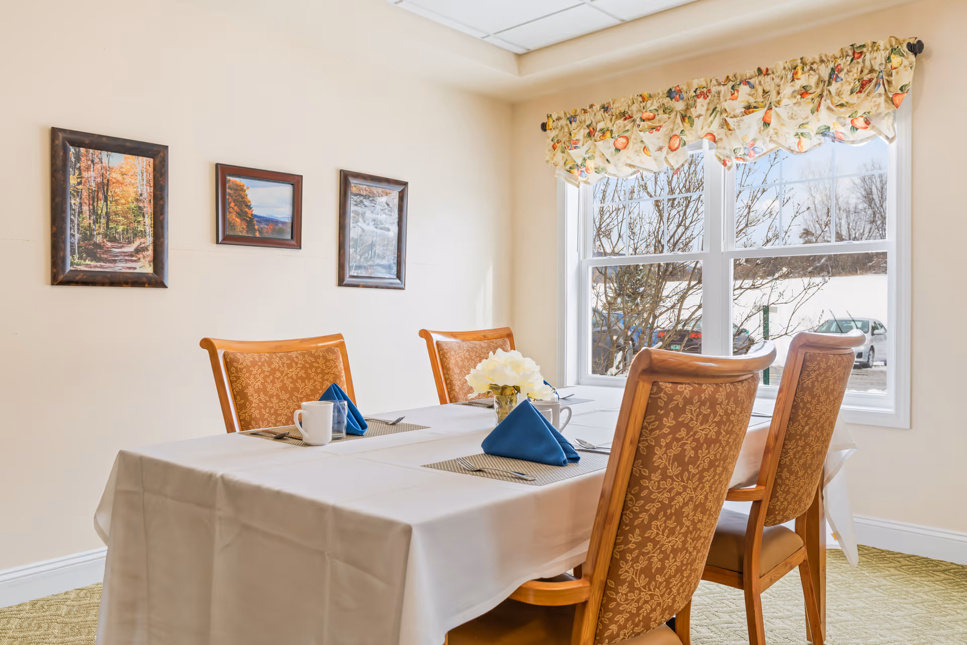 A dining room with a table covered in a white tablecloth set with four place settings, each with a cup, napkin, and utensils. Four wooden chairs with patterned upholstery surround the table. A window with floral valance lets in natural light, and three framed landscape pictures hang on the wall.