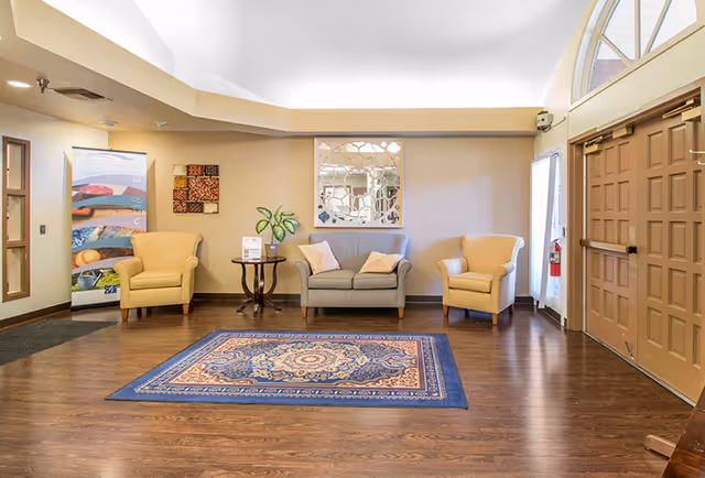 Bright lobby seating area with two yellow armchairs and a gray loveseat arranged around a patterned rug near double entry doors.