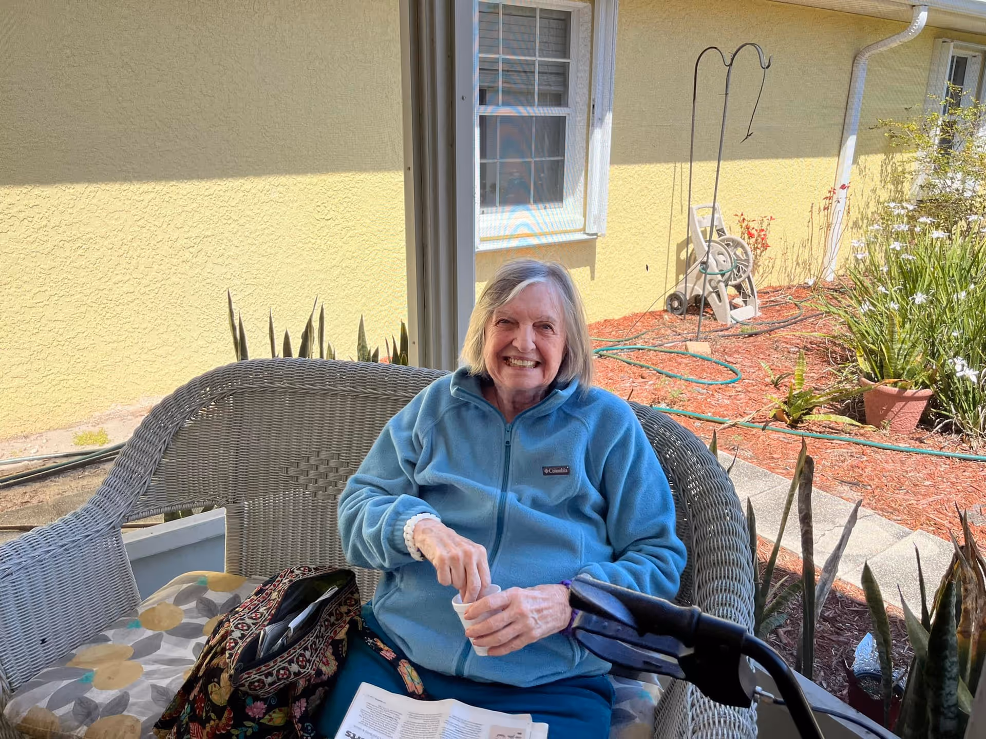 An elderly woman wearing a blue jacket is sitting on a wicker bench outdoors, smiling at the camera. She is holding a small cup and has a walker in front of her. There is a garden with plants and a yellow building wall in the background.