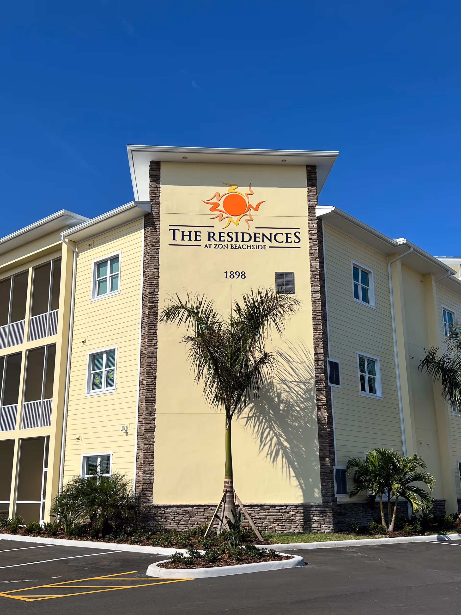 Exterior view of a yellow three-story building with stone accents and a palm tree in front. The building has a sign that reads 'The Residences at Zon Beachside' with a sun logo above it and the number 1898 below. The sky is clear and blue.