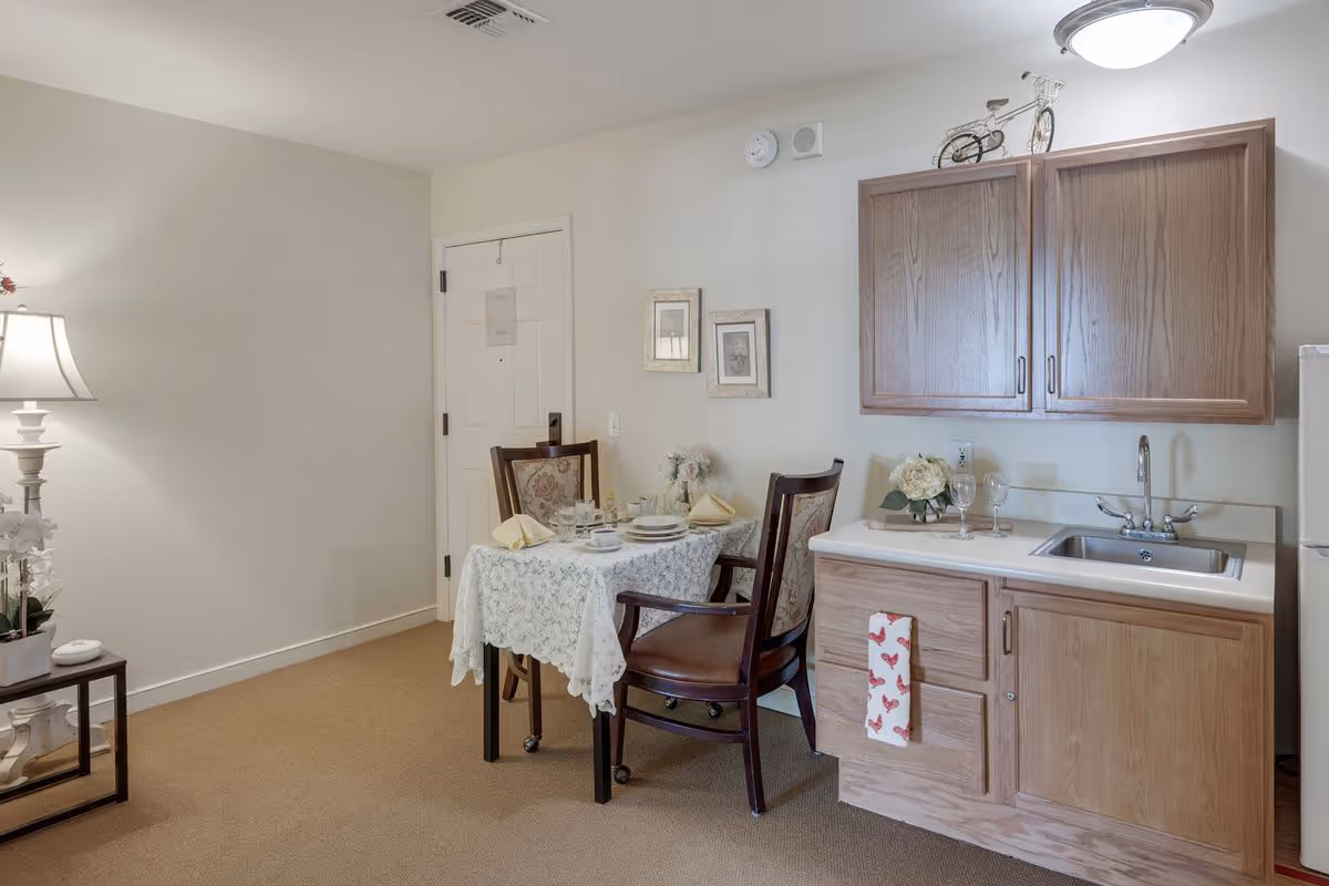 A small dining area with a table covered by a white lace tablecloth set for two, featuring plates, cups, and napkins. Two wooden chairs with floral upholstery are placed at the table. To the right, there is a kitchenette with wooden cabinets, a sink, and a small refrigerator. The room has beige carpet, white walls, a lamp on a side table with flowers, and two framed pictures on the wall.
