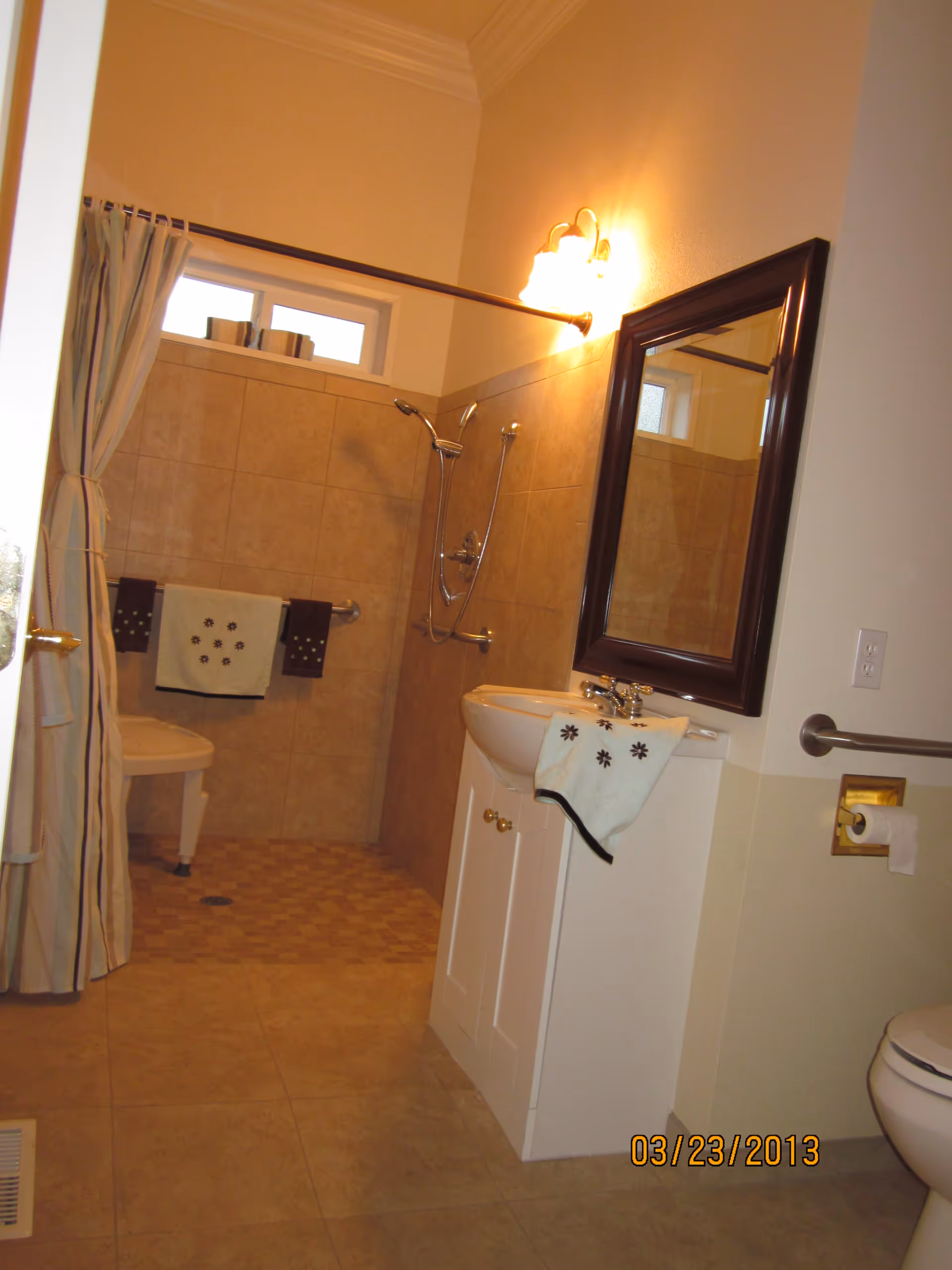 A bathroom with beige tiled walls and floor, featuring a walk-in shower with a shower chair and grab bars. There is a white sink cabinet with a mirror above it and a towel hanging on the sink. A toilet paper holder is mounted on the wall near the toilet, and a shower curtain is partially drawn to the side.
