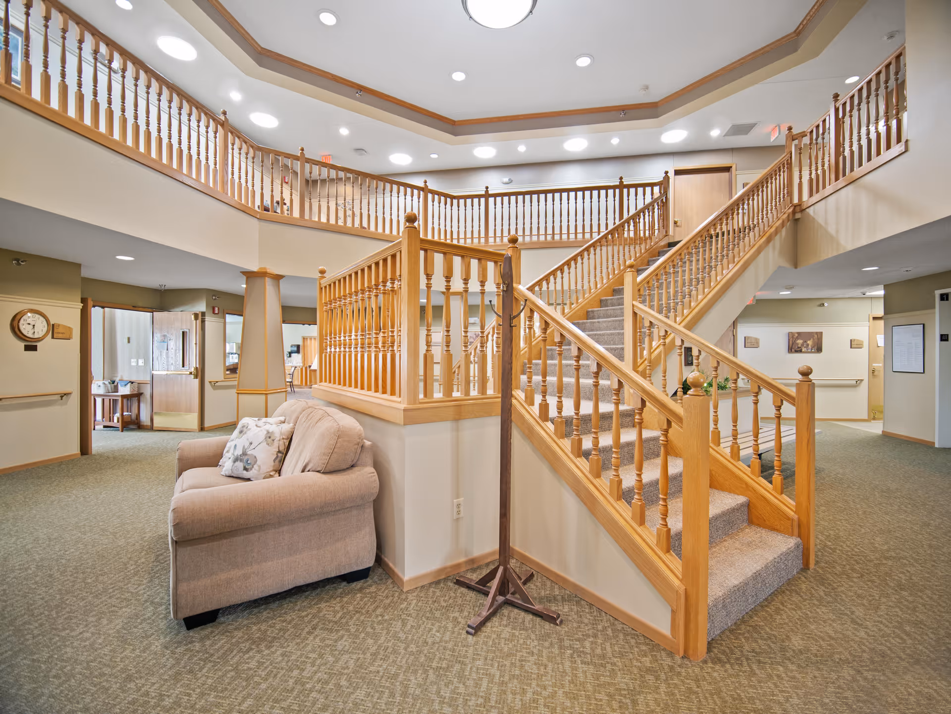 Spacious senior living facility interior lobby with a central wooden staircase, carpeting, and a cushioned armchair.