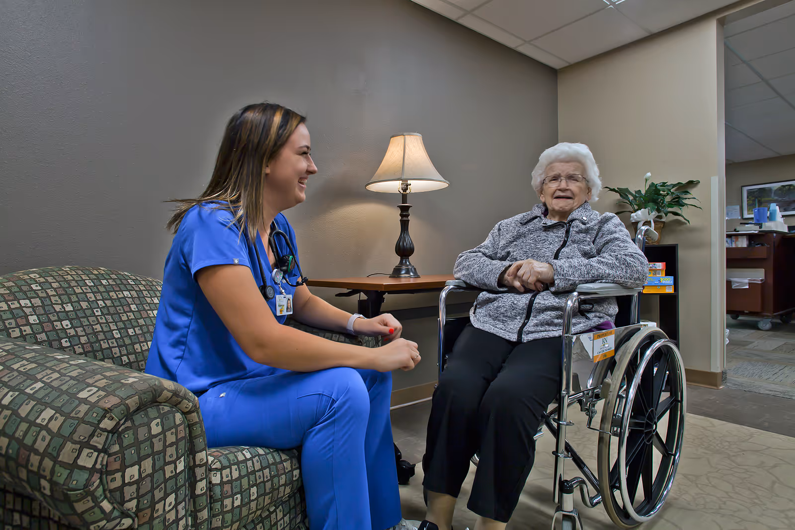A caregiver in blue scrubs chats with an elderly woman seated in a wheelchair in a facility lounge.