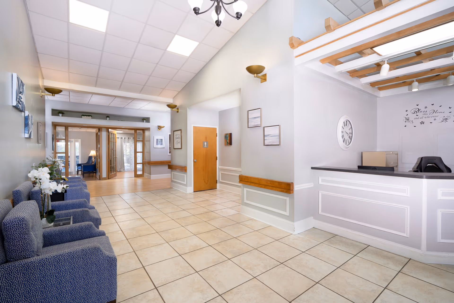 A bright and clean reception area of a senior living facility with a tiled floor, a white reception desk on the right, blue upholstered chairs lined up on the left, and a hallway leading to other rooms. The walls are decorated with framed pictures, and there is a wooden door with a restroom sign. The ceiling has recessed lighting and a chandelier.