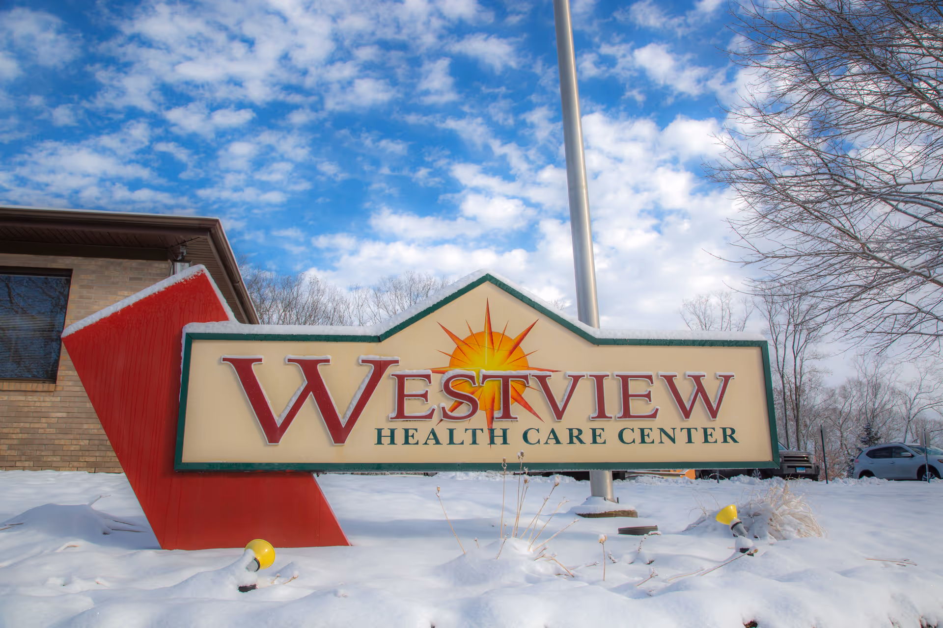 Outdoor view of a large sign for Westview Health Care Center surrounded by snow, with a partly cloudy blue sky and bare trees in the background.