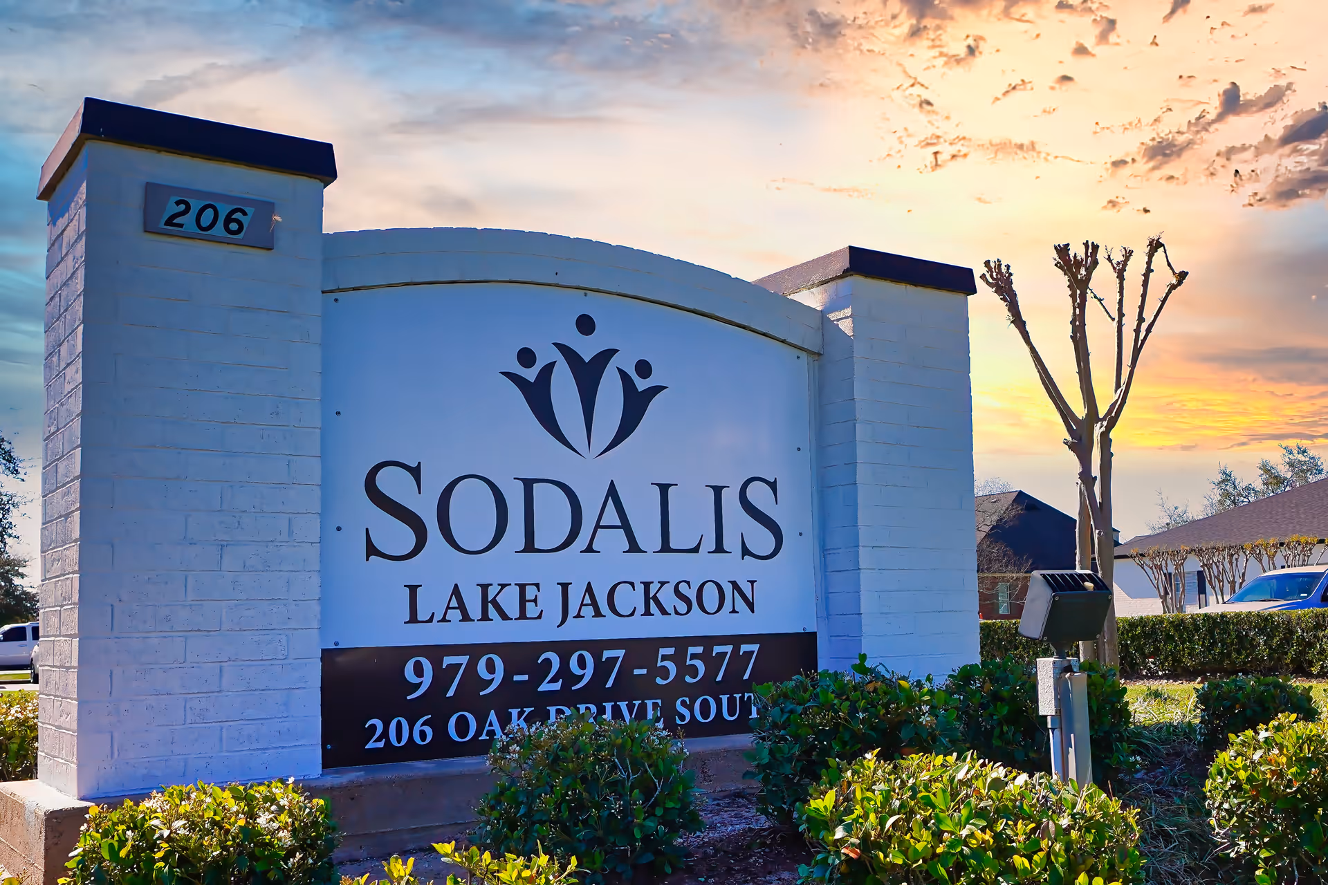Outdoor entrance sign for Sodalis Lake Jackson facility with address 206 Oak Drive South and phone number 979-297-5577, surrounded by bushes and trees under a partly cloudy sky at sunset.