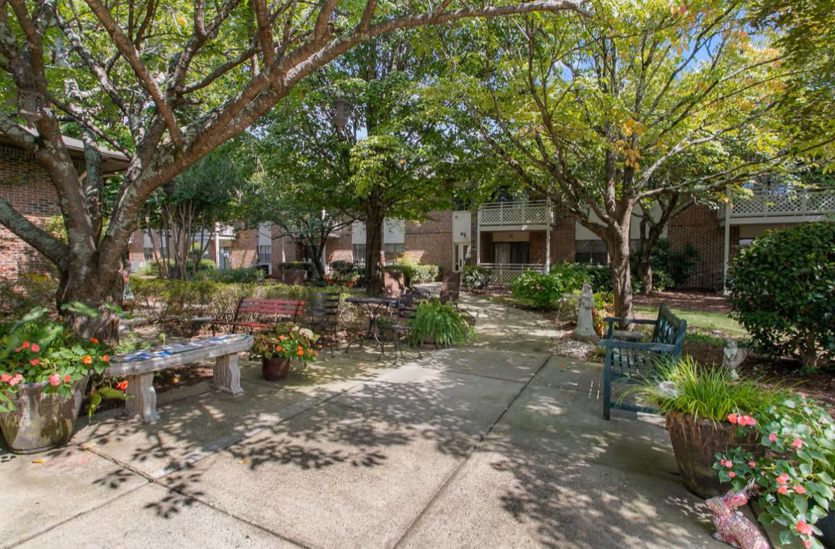 A peaceful outdoor courtyard area at Mount Vernon Village featuring paved walkways, benches, potted plants with flowers, and mature trees providing shade. The background shows brick buildings with balconies and windows.