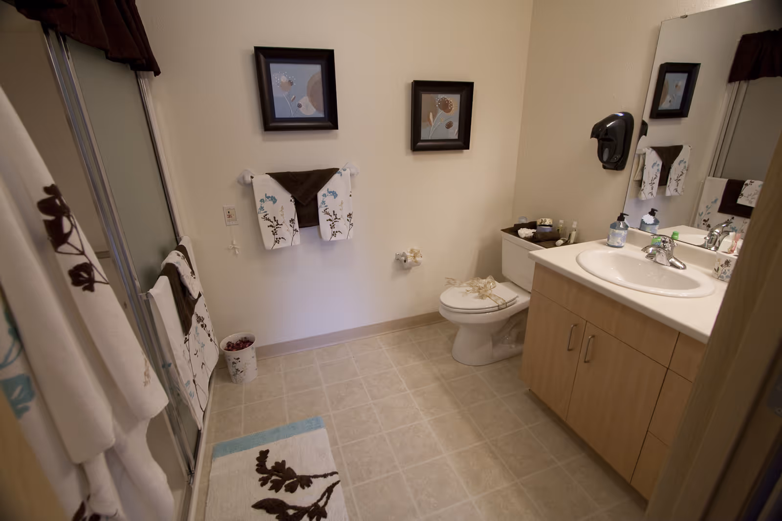 A clean and well-decorated bathroom with a toilet, sink with countertop, large mirror, and a shower with sliding glass doors. The bathroom features beige tiled flooring, light-colored walls, floral-patterned towels, two framed floral artworks on the wall, and a small waste bin.