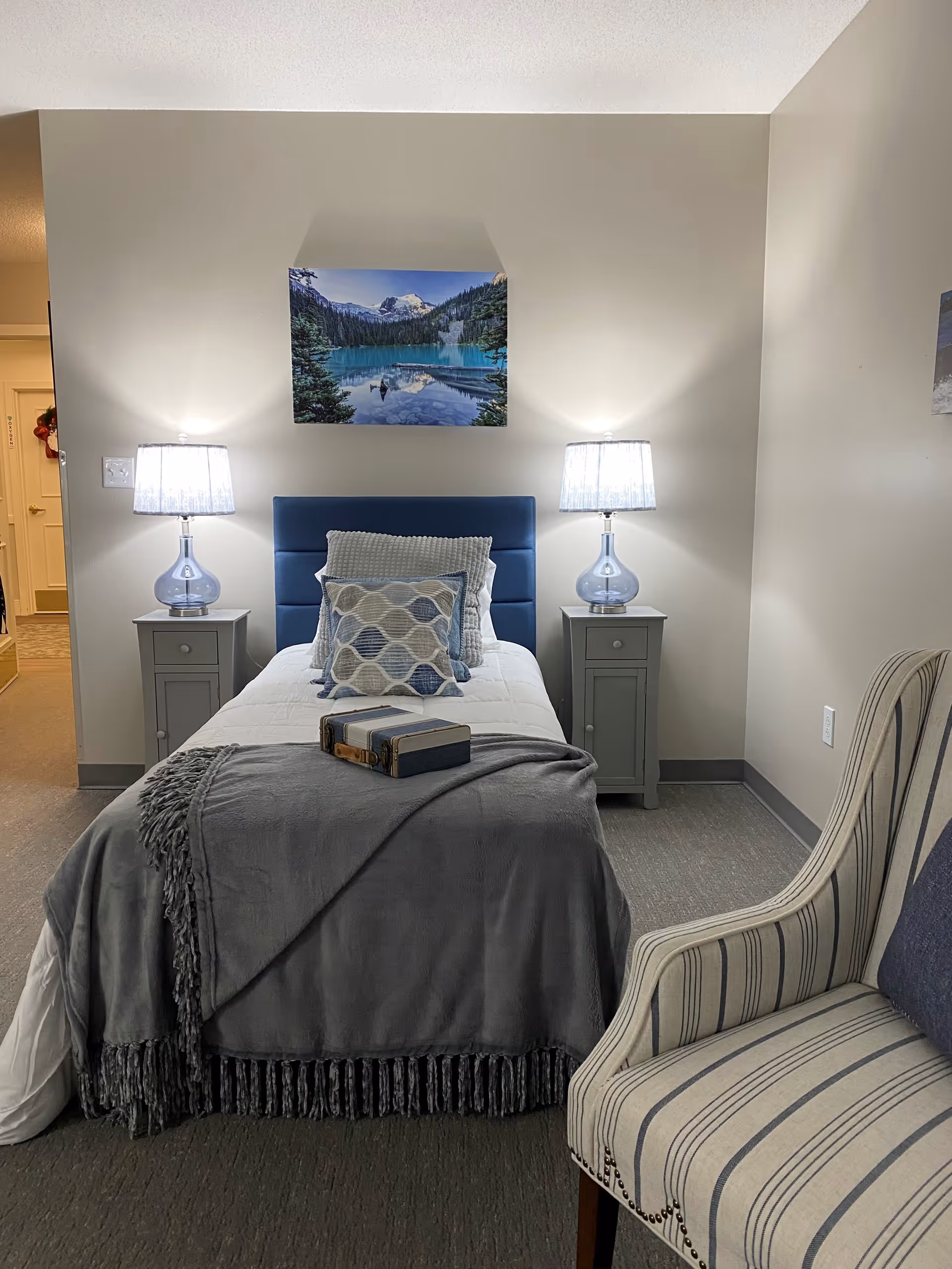 A neatly made single bed with a blue upholstered headboard, white bedding, and multiple decorative pillows. A gray throw blanket is draped over the foot of the bed with two small books placed on top. On either side of the bed are matching gray nightstands with glass table lamps. Above the bed is a landscape painting of a lake surrounded by trees and mountains. To the right of the bed is a striped upholstered armchair. The room has neutral-colored walls and carpeted flooring.
