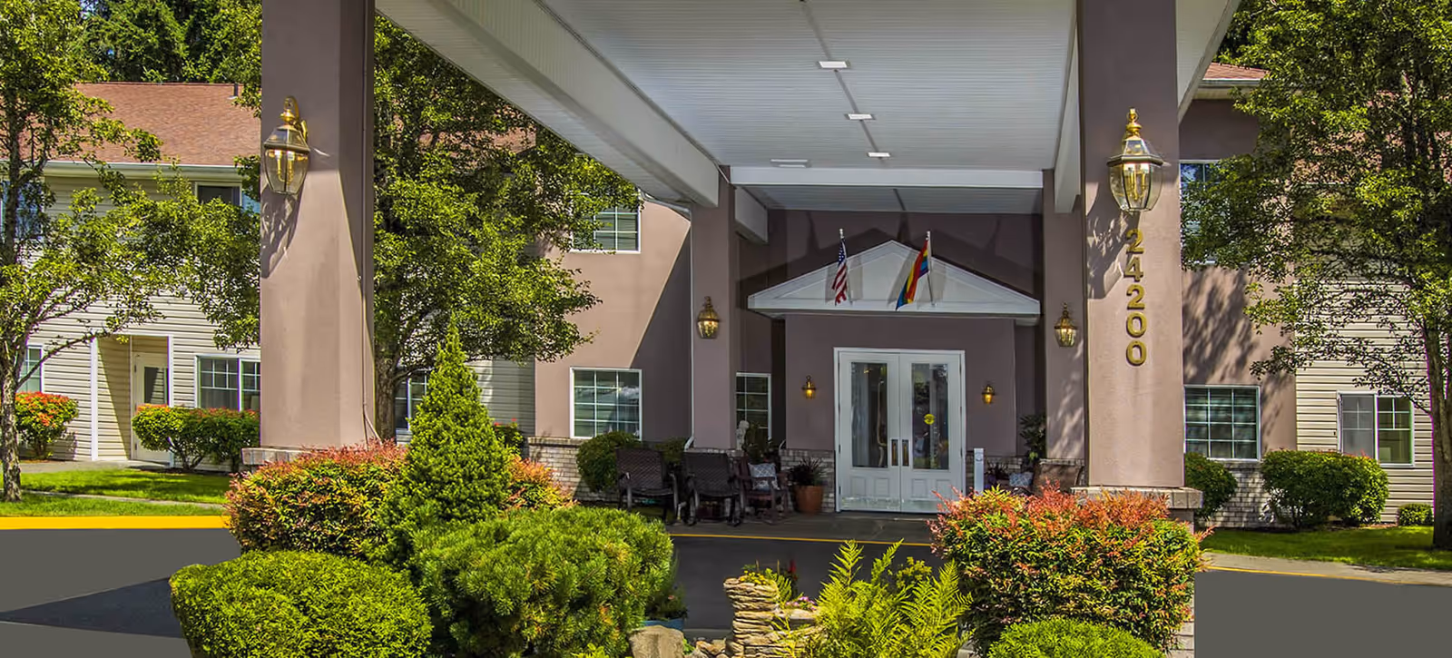 Entrance of a senior living building with a covered porte-cochère, double glass doors, flags above, pillars with address numbers, and landscaped shrubs.