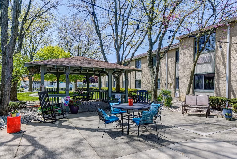 Outdoor patio area at a senior living community with a covered gazebo, metal table and chairs with blue cushions, rocking chairs, benches, potted plants, and string lights hanging from trees. The building is visible in the background with windows and beige brick walls.
