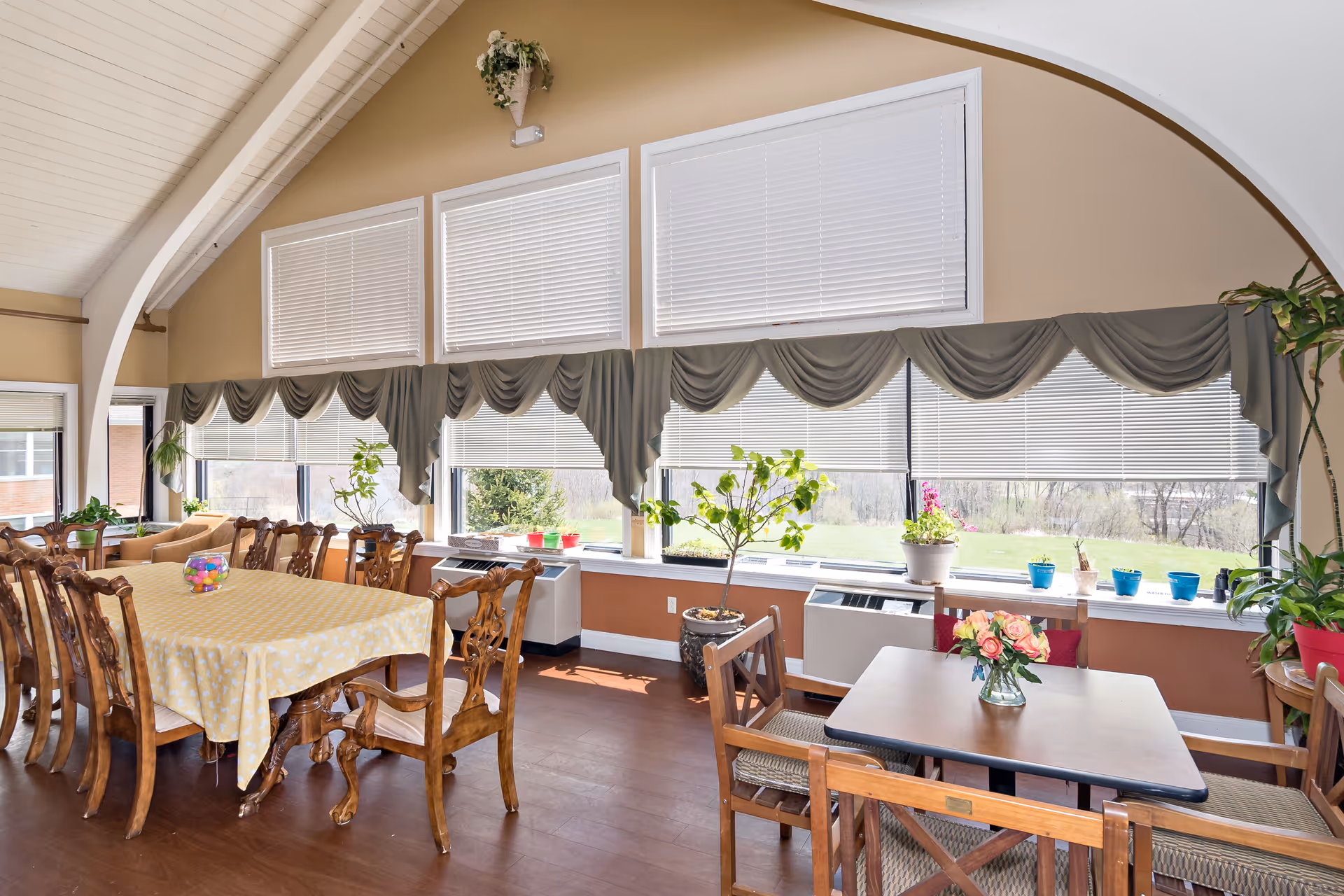 A bright dining room with large windows covered by blinds and decorative valances. The room features a long wooden dining table with a yellow tablecloth and several wooden chairs, as well as a smaller table with a flower vase. Various potted plants are placed on the windowsill and around the room, which has a high, vaulted ceiling with white paneling.