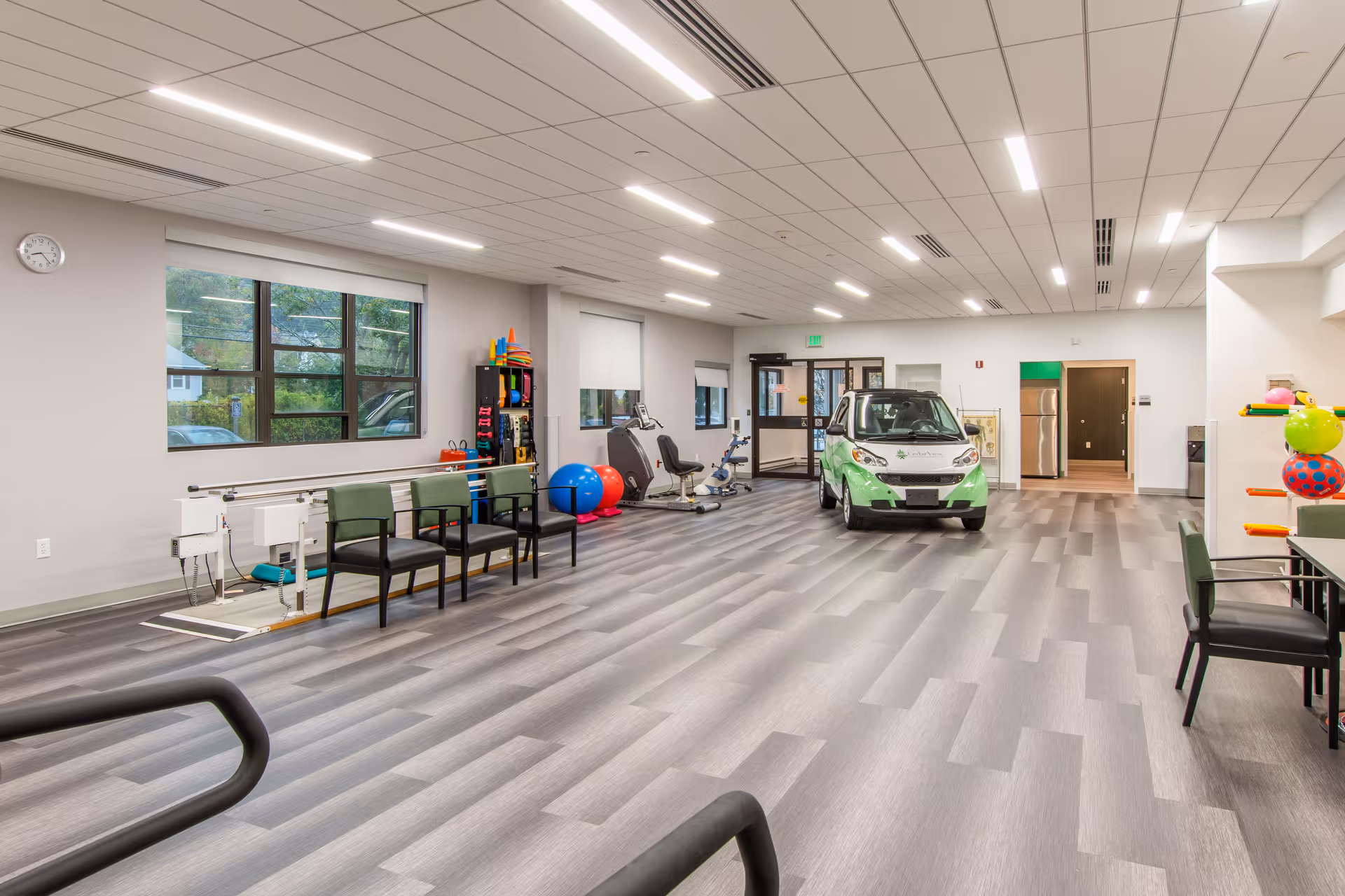 A spacious rehabilitation room with exercise equipment, chairs, therapy balls, and a small green and white car inside. The room has large windows, bright overhead lighting, and a modern gray floor.