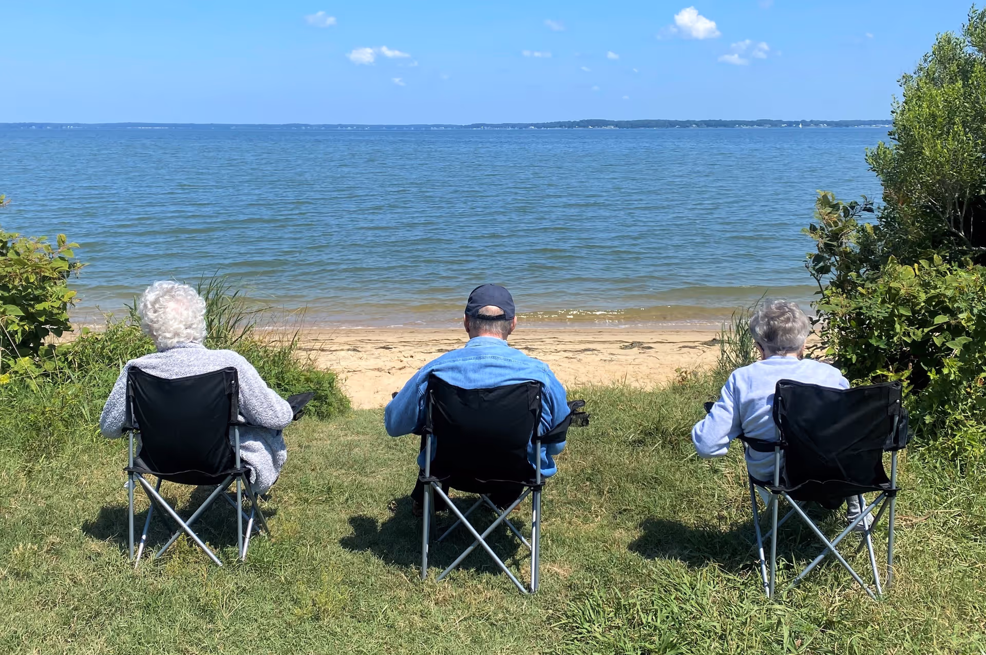 Three elderly people sitting on folding chairs facing a calm body of water with a sandy shore and greenery on either side under a clear blue sky.