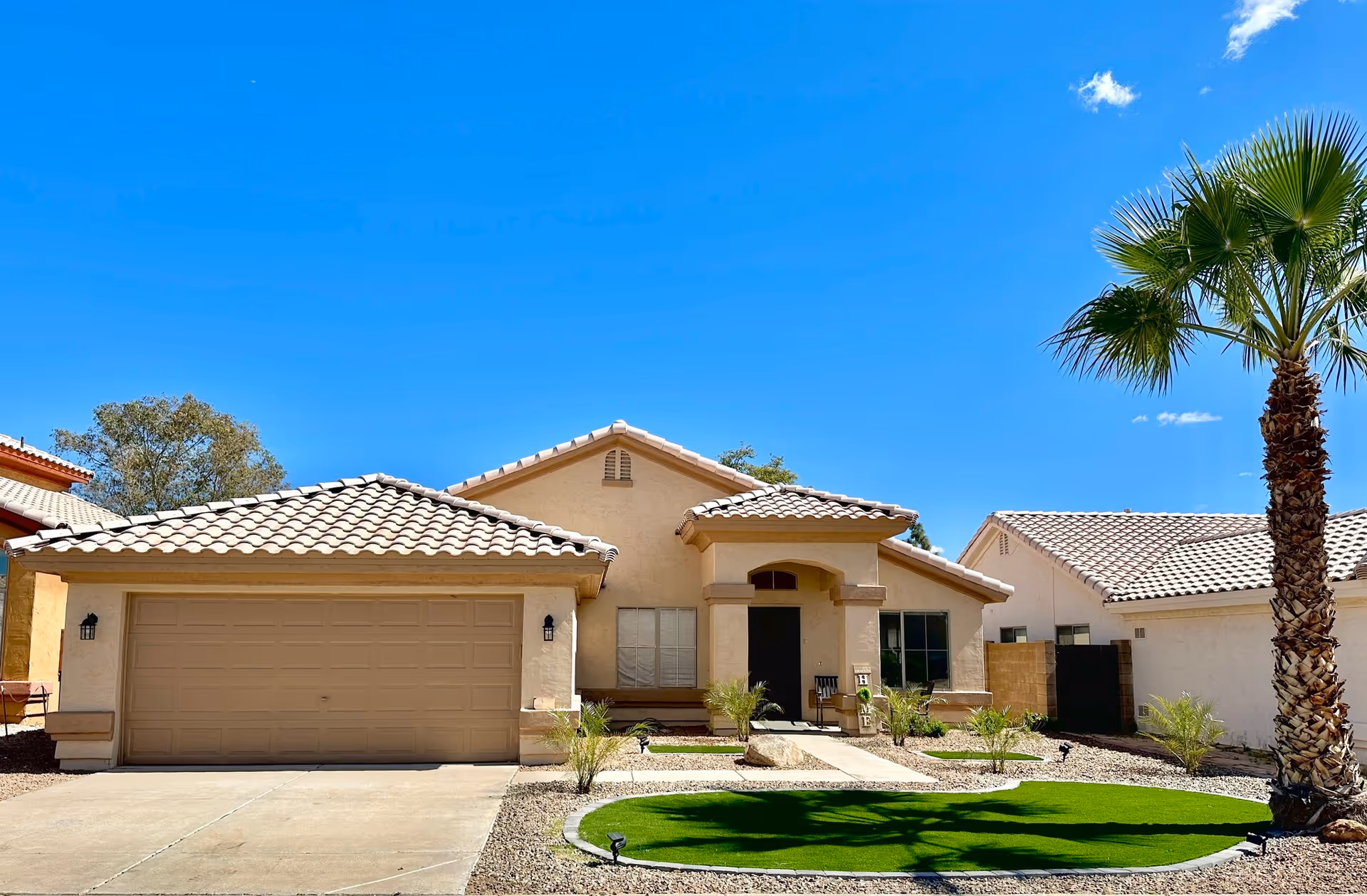 Single-story stucco house with a tiled roof, two-car garage, palm tree and landscaped front yard under a clear blue sky.
