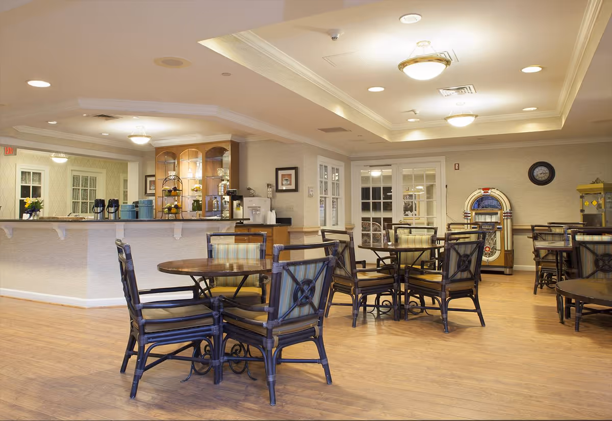 A well-lit dining area with several round wooden tables and cushioned chairs arranged neatly on a wooden floor. In the background, there is a counter with coffee dispensers and a display cabinet with glass doors. A vintage jukebox and a popcorn machine are visible against the far wall. The room has a cozy and inviting atmosphere with ceiling lights and light-colored walls.
