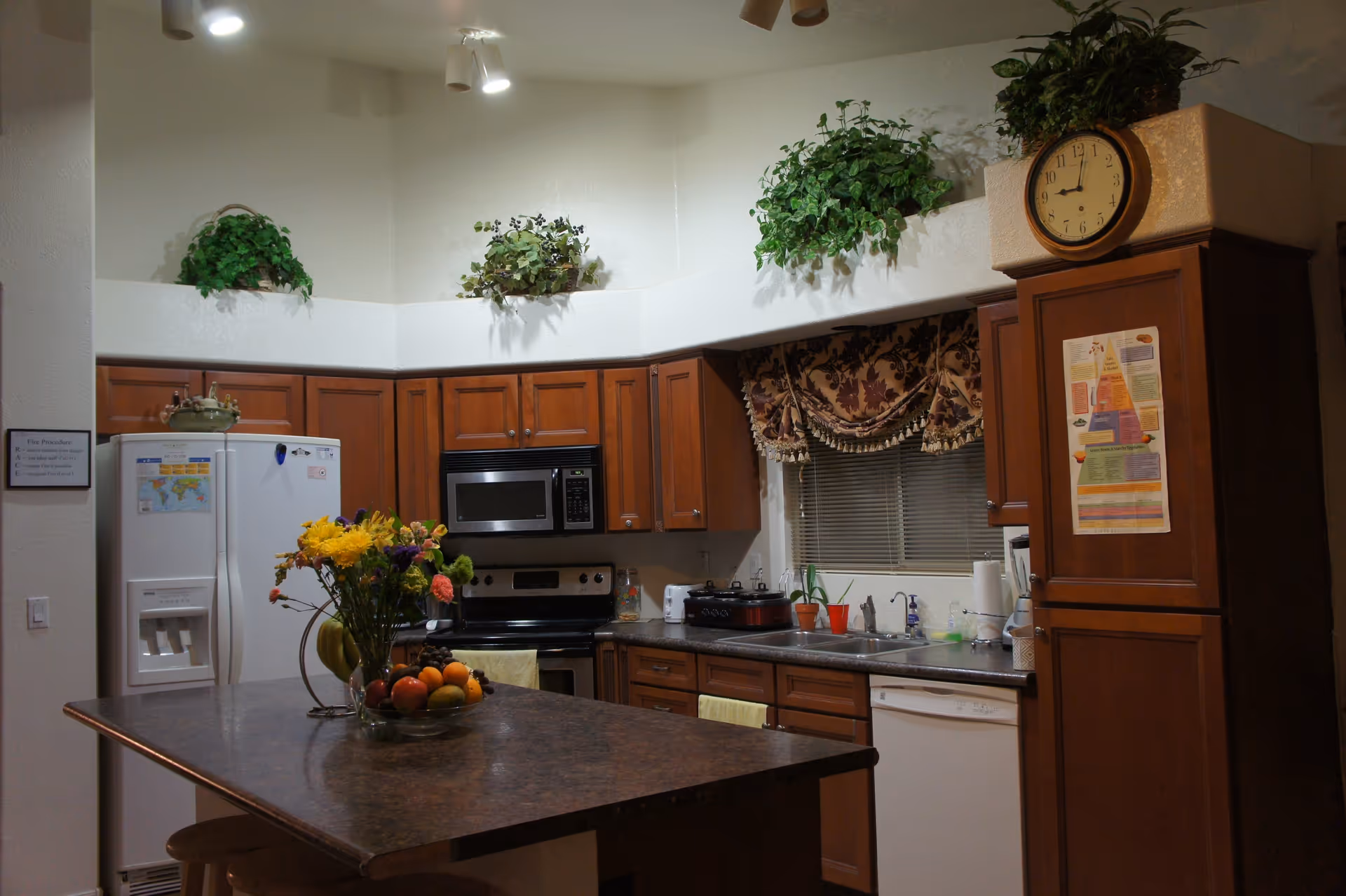 A kitchen with wooden cabinets, a white refrigerator, a microwave above a stove, a dishwasher, and a double sink. There is a kitchen island with a vase of flowers and a bowl of fruit. Above the cabinets are decorative green plants, and a clock is mounted on the wall above a cabinet. A window with a floral valance is above the sink.