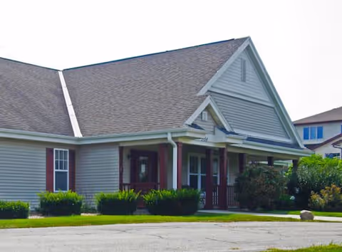 Exterior view of a single-story building with gray siding, a gabled roof, and a covered porch supported by columns. There are bushes and greenery around the building, and a paved driveway or parking area in front.