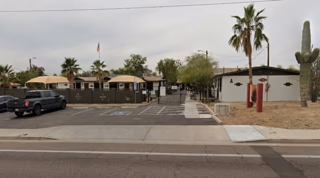 Front exterior view of The Park at 7th Avenue facility showing a gated entrance, parking spaces including a handicapped spot, palm trees, a large cactus, and single-story buildings under a cloudy sky.