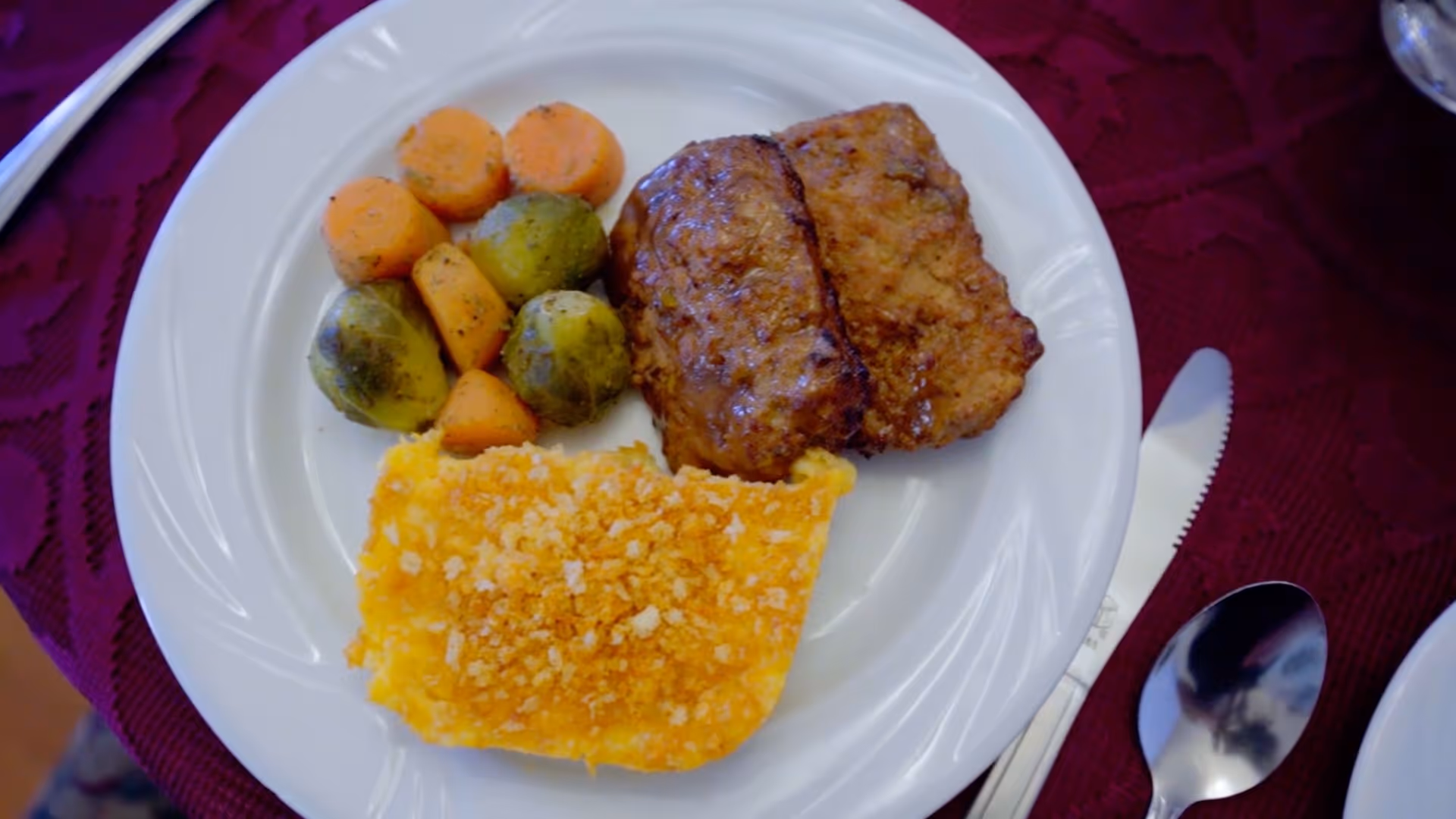 A white plate with a serving of cooked carrots, Brussels sprouts, two pieces of meatloaf, and a portion of baked macaroni and cheese on a maroon tablecloth. A knife and spoon are placed to the right of the plate.